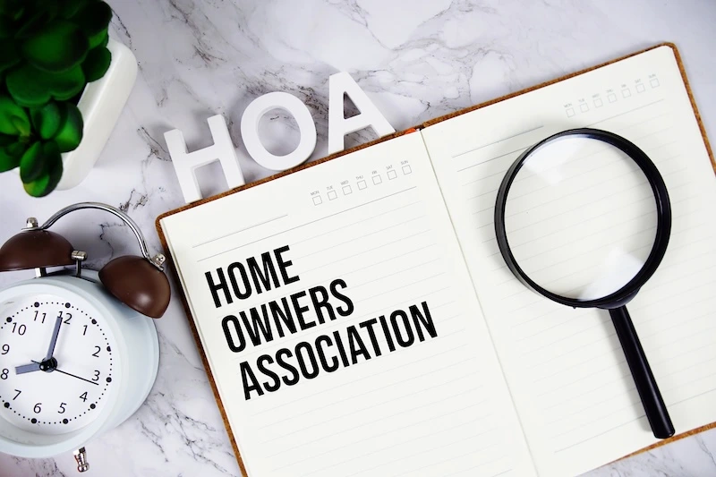 An open notebook on a marble surface displays the words Home Owners Association. A magnifying glass rests on the notebook, with an alarm clock, glasses, and white letters spelling HOA nearby.