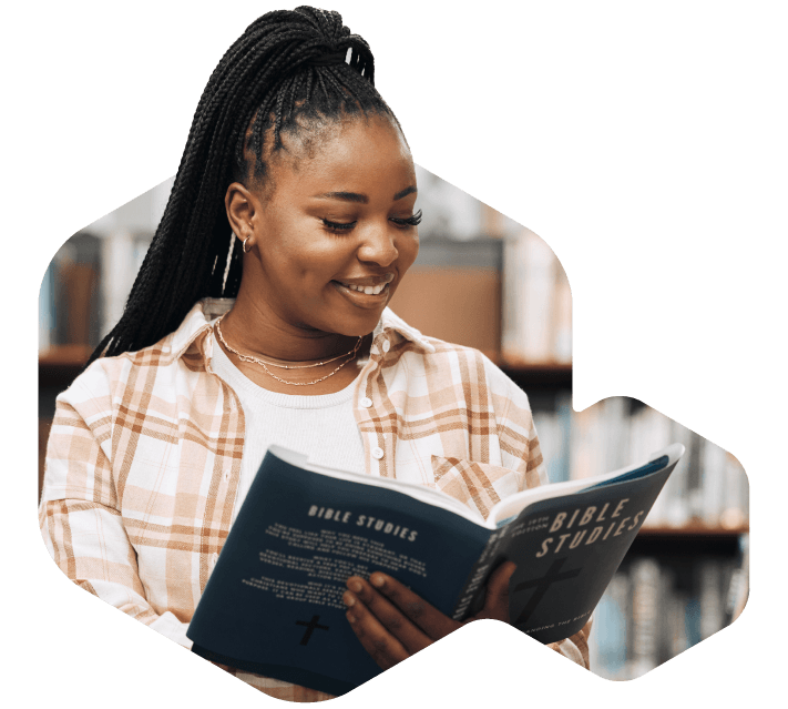 A young woman with braided hair smiles while reading a book in a library setting, possibly researching faith based voting software. She wears a plaid shirt over a white top and layered necklaces.