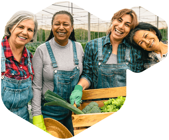 Four women in overalls and gloves smile together while harvesting vegetables in a greenhouse. They stand close to each other, holding fresh produce in a wooden crate.
