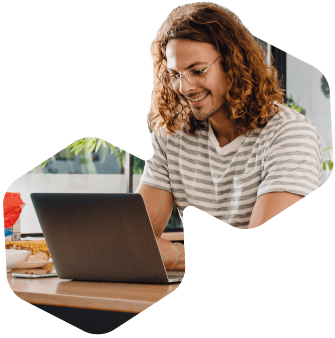 A person with long curly hair and glasses, wearing a striped shirt, smiles while using an HOA voting tool on a laptop at a wooden table in a bright, indoor setting.