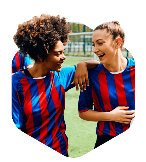 Two young women in blue and red striped soccer jerseys stand on a field, laughing together after an online team voting session, with one resting her arm on the other's shoulder.