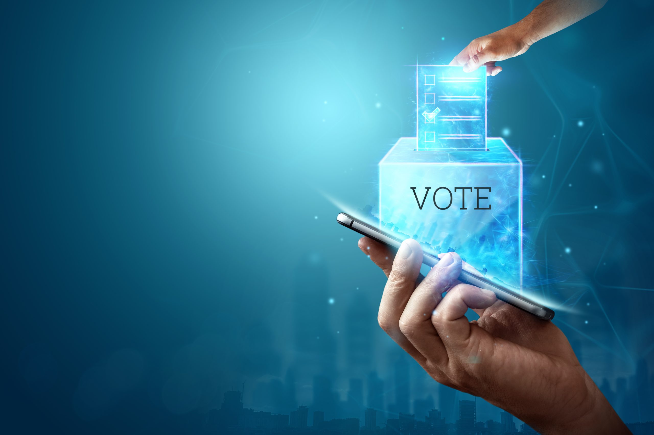 A hand holds a smartphone emitting digital light, while another hand places a glowing ballot into a transparent box labeled VOTE, symbolizing online or electronic voting. The background is blue with subtle digital effects.