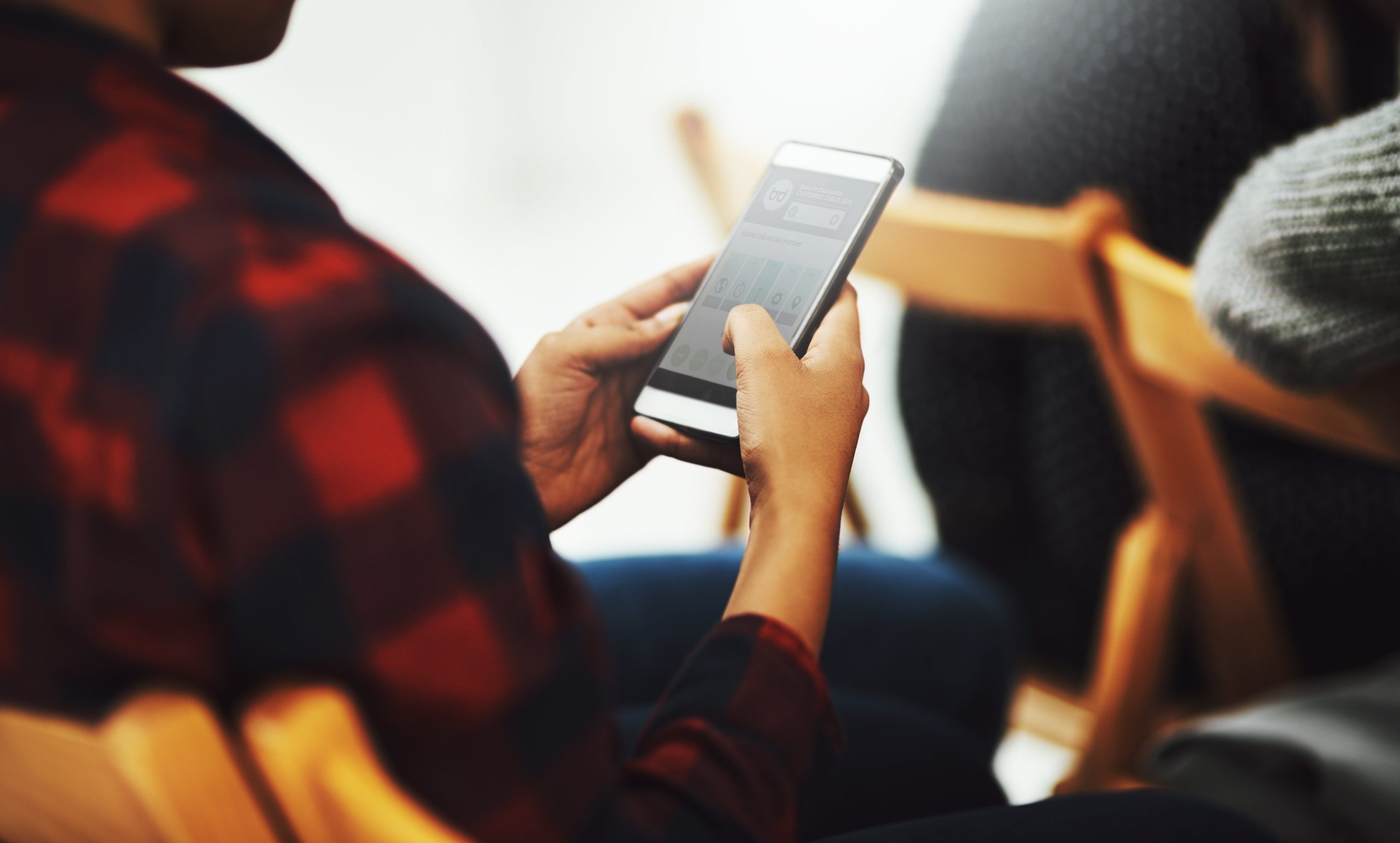 A person wearing a plaid shirt is sitting and using a smartphone, tapping on the screen. Other people seated nearby are slightly out of focus in the background.